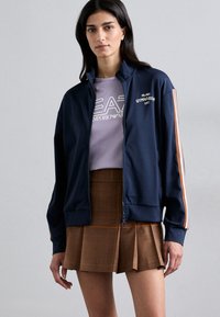 Navy zip-up jacket with high collar, white embroidered text, and orange stripes on sleeves. Paired with a light purple t-shirt and plaid brown skirt.