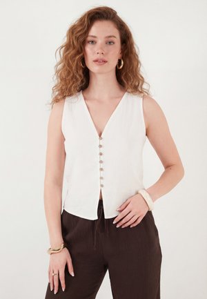 Göndör hajú nő, aki ujjatlan, fehér, gombos felsőt és sötét texturált nadrágot visel, kiegészítve karika fülbevalókkal és karkötőkkel.