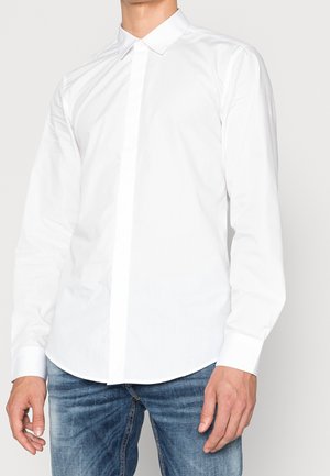 Mężczyzna w białej koszuli na długi rękaw z kołnierzykiem i niebieskich dżinsach, stojący na jednolitym tle.