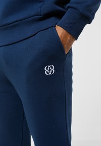 Rankoje mėlynos spalvos sportinių kelnaičių kišenėje, ant šlaunies yra baltas siuvinėtas abstraktinis logotipas, derinamas su atitinkama mėlyna sportine palaidine.