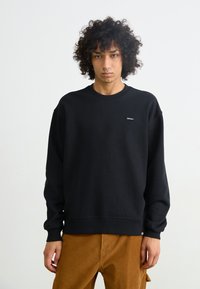 Zwarte sweatshirt van katoen, met een ronde hals, geribbelde manchetten en een klein merkje op de borst. Gekleed met een bruine corduroy broek.