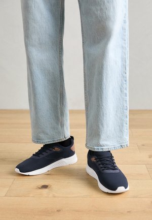 Person trägt hellblaue Jeans und schwarze Turnschuhe mit weißen Sohlen und steht auf einem Holzboden.