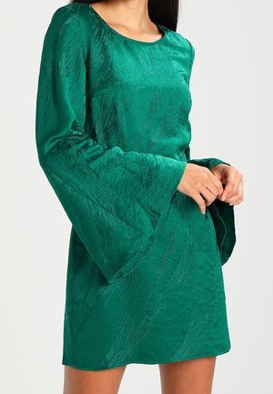 Kvinde iført en skinnende tekstureret smaragdgrøn minikjole med lange ærmer og brede klokkeærmer, stående med hænderne foldet ved taljen.