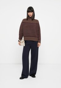 Maglione a righe marroni in maglia con collo alto e vestibilità ampia, abbinato a pantaloni navy a gamba larga. La modella tiene una piccola borsa beige.