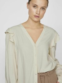 Cremefarvet bluse med flæseærmer, V-udhuling og knaplukning foran. Fremstillet af letvægts tekstureret stof, parret med højtaljede brune bukser.