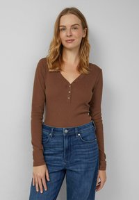 Brązowa bluzka z długim rękawem, dekoltem w serek i ozdobnymi guziczkami, zestawiona z wysokim stanem niebieskich dżinsów o prążkowanej fakturze.