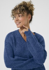 Blauer gestrickter Pullover mit V-Ausschnitt, langen Ärmeln und gerippter Textur. Der Stoff wirkt weich und gemütlich und zeigt eine entspannte Silhouette.