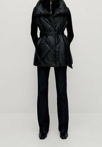 Sort quiltet ærmeløs vest med højt krave, bælte i taljen og en glansfuld finish, kombineret med mørke denim flared bukser og elegante sorte sko.