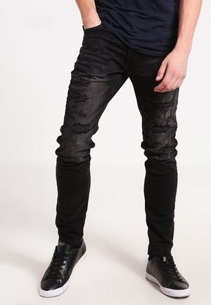 Person trägt schwarze, ausgefranste Skinny-Jeans und schwarze Sneakers mit weißen Sohlen, steht mit einer Hand in der Tasche.
