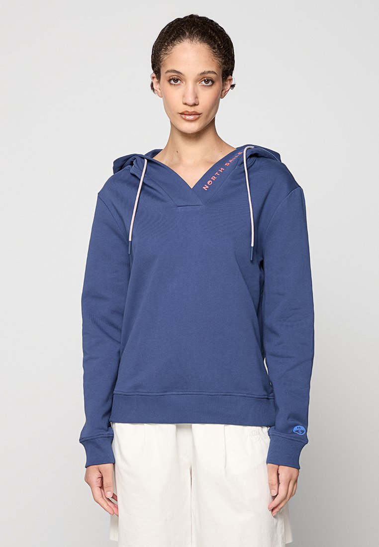 Frau in einem blauen Kapuzenpullover mit Kordeln und weißen Hosen, die vor einem schlichten hellen Hintergrund steht.