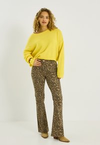 Mujer con cabello rizado, vestida con un suéter amarillo, pantalones de talle alto con estampado de leopardo y botas de tacón beige, posando frente a un fondo liso.