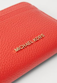 Rdeča teksturirana usnjena denarnica z zlatim logotipom "MICHAEL KORS" in horizontalnim šivanjem na vrhu. Kompaktna oblika.