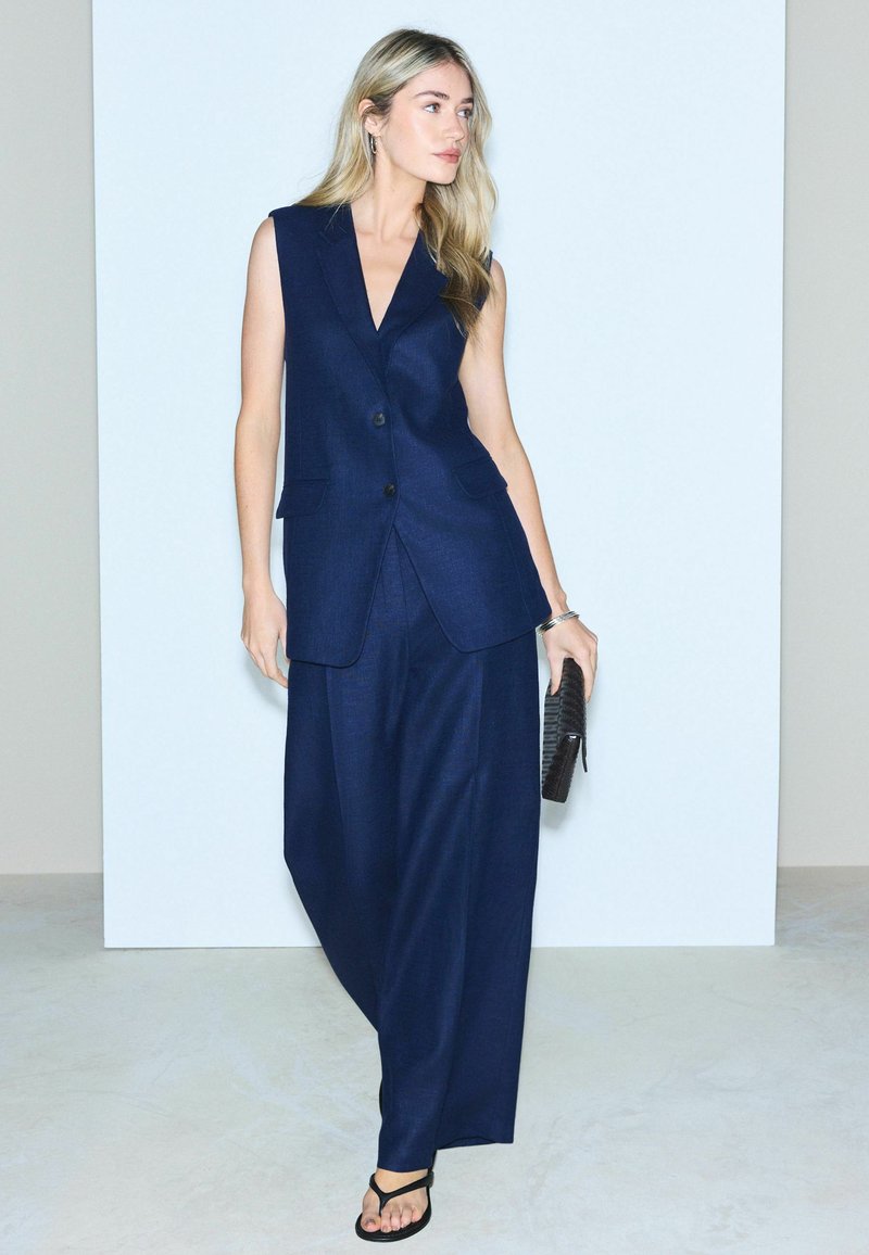 Donna che indossa un completo senza maniche blu navy con pantaloni a gamba larga, tiene una pochette nera, in piedi davanti a uno sfondo chiaro e uniforme.