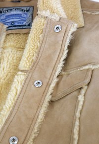 Tannfarbene Lammfelljacke mit einem flauschigen beigen Futter. Sichtbare silberne Knöpfe und gestickte Nahtdetails. Das Etikett zeigt die Größe "M".