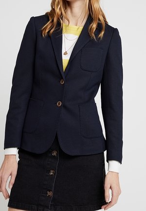Kvinde iført en tætsiddende marineblå blazer med to forlommer, brune knapper, lagdelt over en stribet gul-hvid top og et sort knapbesat skørt.