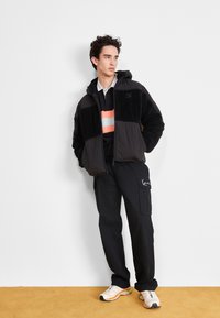 Veste en polaire noire et matelassée avec capuche, portée sur une chemise colorée et un pantalon cargo noir, assortie de baskets multicolores.