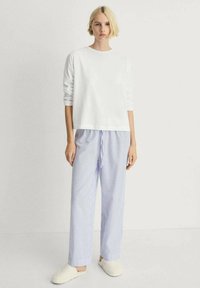 White long-sleeve top, blue and white striped pajama pants with a drawstring; model wearing beige slippers, standing against a neutral background.