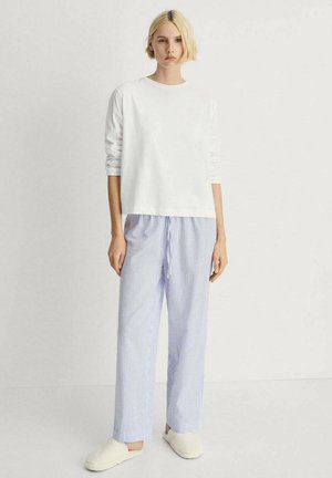 Witte lange mouwen top, blauw-witte gestreepte pyjamabroek met een trekkoord; model draagt beige slippers en staat tegen een neutrale achtergrond.