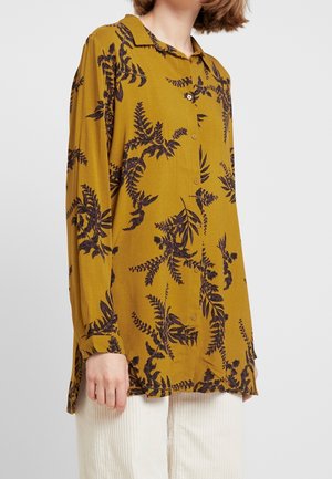 Person iført en knallergul skjorte med sorte blomsterblade og hvide ribbede bukser, beskåret ved hals og hofter.