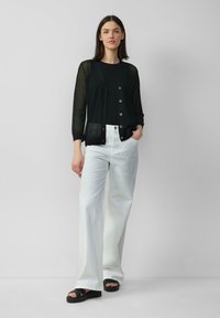 Schwarze Strickjacke mit offener Maschenstruktur und silbernen Knöpfen, kombiniert mit einem schwarzen Oberteil und weißen, weit geschnittenen Jeans. Schwarze Plattformsandalen runden den Look ab.
