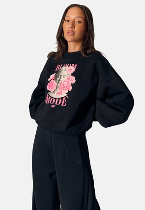 Kobieta w czarnej bluzie z różowym graficznym motywem kwiatowej rzeźby i napisem „Bloom Mode”, w luźnych czarnych spodniach, stojąca na białym tle.