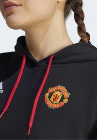 Sweat à capuche noir avec cordons rouges et logo brodé de Manchester United sur la poitrine. Tissu : mélange de coton, texture lisse.