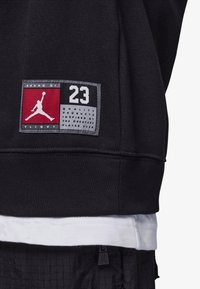 Felpa nera con etichetta rossa e grigia che mostra il logo Jumpman. Sotto si intravede una maglietta bianca. Dettagli testuali inclusi sull'etichetta.