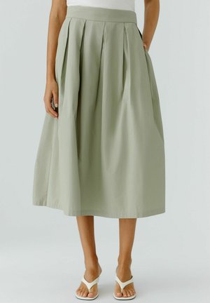 Femme portant une jupe midi plissée taille haute vert sauge clair avec poches et des sandales blanches à talons ouverts, debout devant un fond uni.