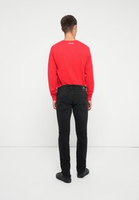 Rdeča majica z dolgimi rokavi in okroglim izrezom, z belim napisom na hrbtu, v kombinaciji s črnimi slim-fit jeans hlačami in črnimi čevlji.