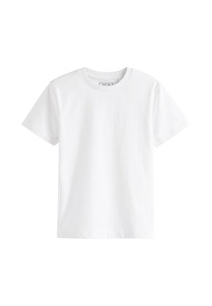 Junge mit lockigem Haar steht und schaut nach rechts, trägt ein schlichtes weißes kurzärmeliges Shirt, schwarze Hose und graue Turnschuhe vor beigem Hintergrund.