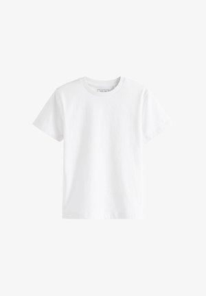 Dreng med krøllet hår står og kigger mod højre iført en ensfarvet hvid kortærmet t-shirt, sorte bukser og grå sneakers mod en beige baggrund.