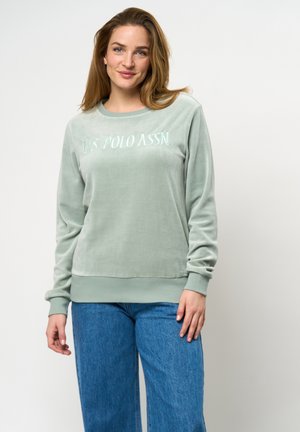 Mintgroene velours sweatshirt met borduursel van U.S. Polo Assn. Kenmerken zijn lange mouwen en een ronde halslijn, gecombineerd met blauwe wijde jeans.
