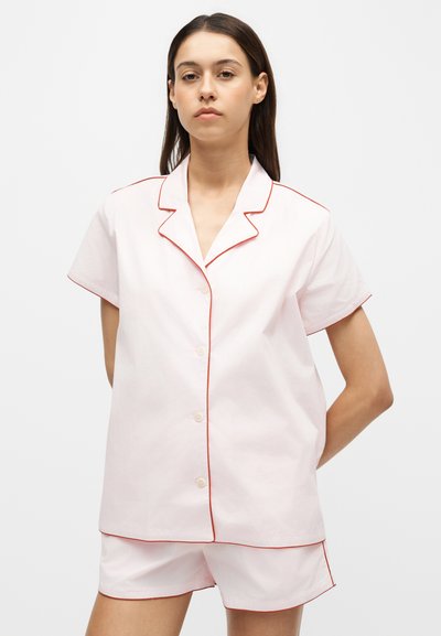 Femme portant un haut de pyjama rose clair à manches courtes boutonné et un short assorti avec un passepoil rouge, debout les mains derrière le dos.