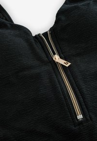 Zbliżenie na czarną, teksturowaną sukienkę z złotym zamkiem błyskawicznym częściowo rozpiętym z przodu.