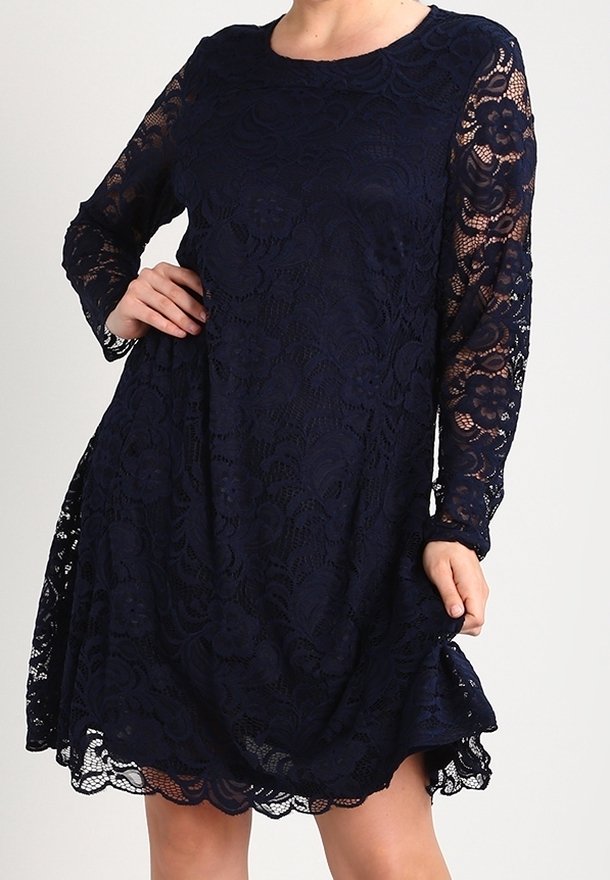 Femme portant une robe en dentelle bleu marine à manches longues avec des motifs floraux, tenant légèrement la jupe d'une main.