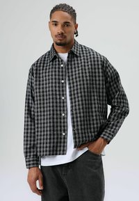 Zwart-grijs geruit overhemd met een knoopsluiting aan de voorkant, lange mouwen en een ontspannen pasvorm, gedragen over een wit T-shirt. Getextureerde stof.