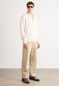 Beige gebreide sweater met een kraag, lange mouwen; gecombineerd met crèmekleurige op maat gemaakte broek en donkerbruine veterschoenen; draagt een zonnebril.