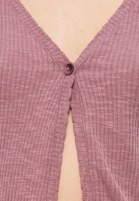 Μοβ πλεκτή ζακέτα με V-λαιμόκοψη, ριμπ υφή και ένα στρογγυλό κουμπί κλεισίματος στο στήθος.