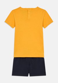 Κίτρινο βαμβακερό T-shirt με κοντά μανίκια και κλείσιμο με κουμπί στο πίσω μέρος, συνδυασμένο με τσέπες μπλε βαμβακερά σορτς. Απλός σχεδιασμός, μαλακή υφή.