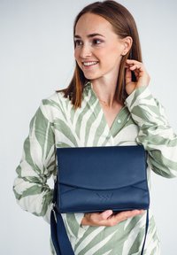 Námořnická kožená kabelka přes rameno s klopou a nenápadným logem. Hladký povrch a nastavitelný popruh pro různorodé možnosti nošení.