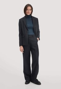 Zwarte op maat gemaakte suit met een gestructureerde blazer, wijde broekspijpen en een donkere coltrui. Glad materiaal met een klassieke pasvorm. Zwarte loafers maken de look compleet.