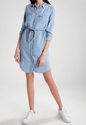 Femme portant une robe chemise en denim bleu clair avec des manches retroussées, cintrée à la taille avec une ceinture marron, et des baskets blanches avec des touches rouges.