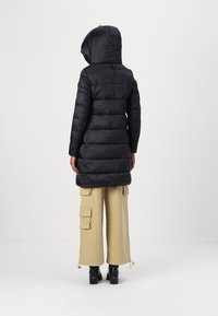 Black puffer coat with a hood, featuring a quilted design and a mid-length cut, worn over beige cargo pants and black heeled boots.