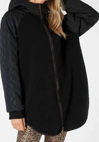 Giacca in pile nera con maniche trapuntate nere, zip frontale e cappuccio. Presenta un orlo arrotondato e polsini a coste. Collant con stampa leopardata sotto.