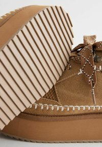 Sneaker in suede marrone con suola in gomma testurizzata e rigata. Presenta dettagli cuciti e un laccio intrecciato con un motivo a diamante.