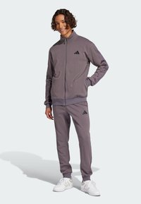 Šedý zipovací teplákový komplet vyrobený z měkkého materiálu, který se vyznačuje uvolněným střihem, žebrovanými manžetami a černými logovými akcenty na bundě a kalhotách.