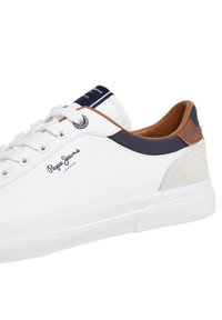 Hvite skinnsneakers med grå og brune detaljer, logodetalj på siden, rund tå og mønstret gummisåle for grep.