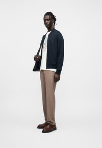 Μπλε σκούρο φούτερ με κουμπιά τύπου σνάπ, λευκό μπλουζάκι με τυπωμένο σχέδιο, μπεζ παντελόνι και καφέ παπούτσια, συνδυασμένα με μαύρη τσάντα ώμου.