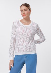 Λευκό κορδονέ μπλούζα με λουλούδια, μακριά μανίκια, στρογγυλή λαιμόκοψη και ριμπ μανσέτες και τελείωμα. Συνδυάζεται με ανοιχτό μπλε τζιν.