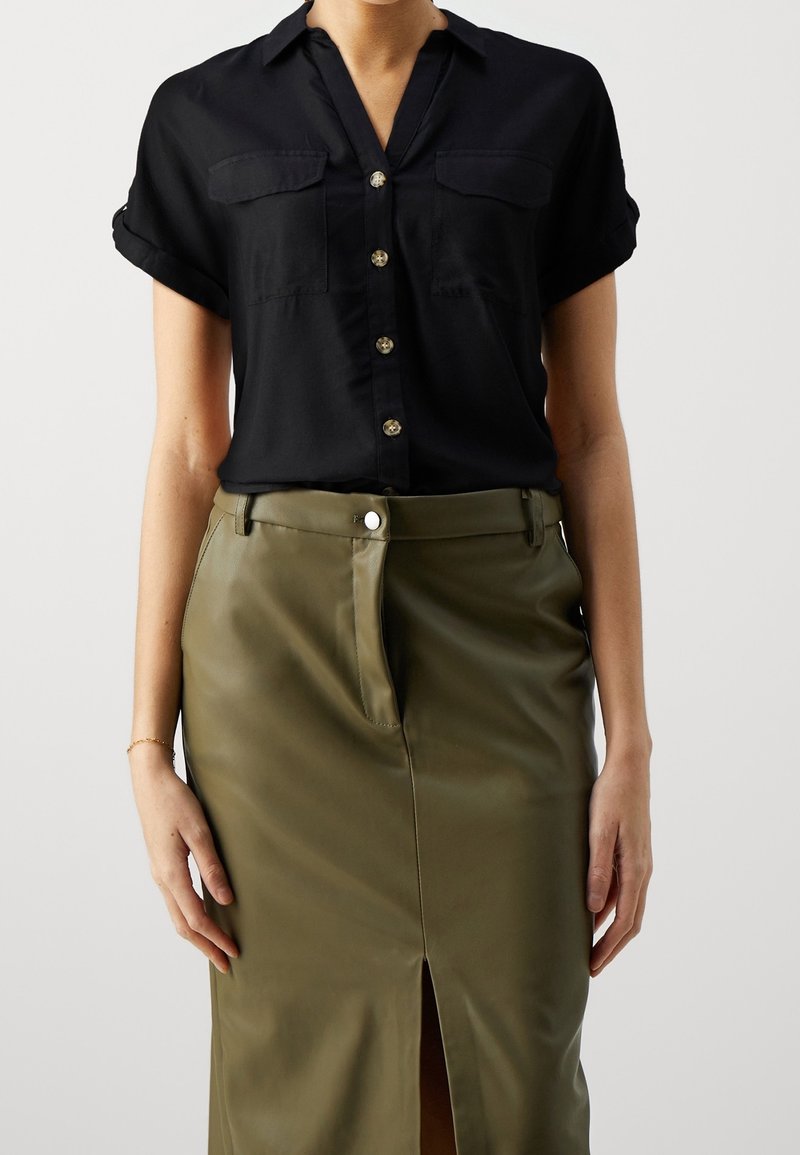 Camisa de botones negra de manga corta con dos bolsillos en el pecho, combinada con una falda de cuero verde oliva de talle alto que presenta una abertura frontal.